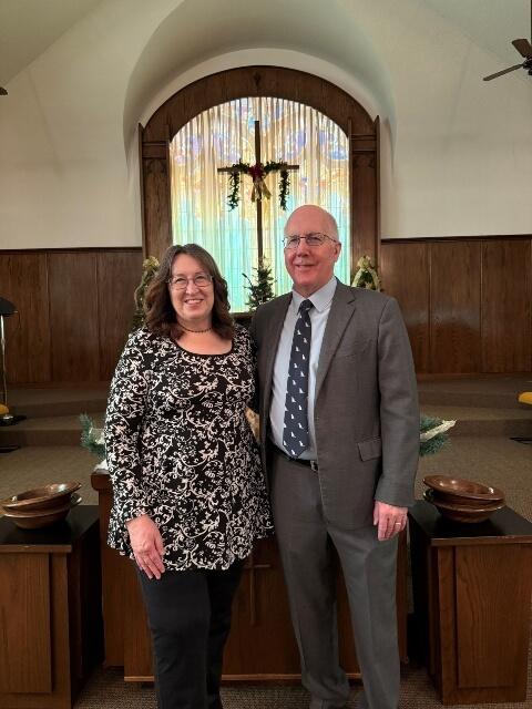 Pastor Bruce and Linda Wells standing at the front of the sanctuary. In the background you can see a cross and stained glass window.